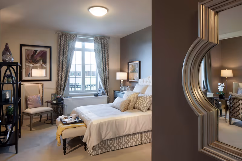 A cozy and well-decorated bedroom in a senior living facility featuring a neatly made bed with multiple pillows, a cushioned bench at the foot of the bed with a tray, a chair with a pillow, a window with patterned curtains, framed artwork on the walls, a side table with a lamp, and a decorative mirror reflecting part of the room.