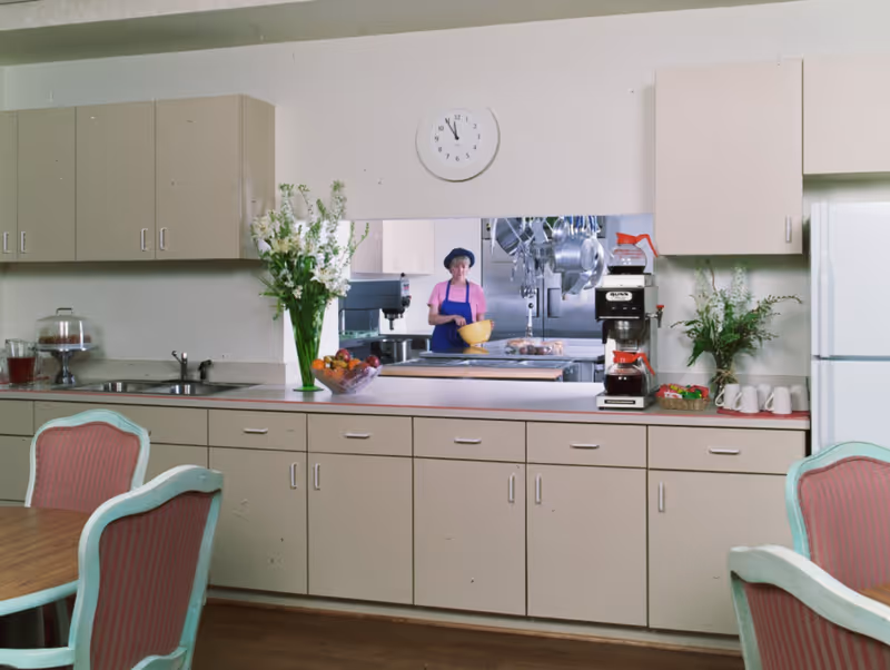 Interior view of a kitchen area with beige cabinets and drawers, a sink, a coffee maker, a bowl of fruit, and flower vases on the countertop. A window opening shows a person in a pink shirt and blue apron holding a yellow bowl in a commercial kitchen. There are pink cushioned chairs around a wooden table in the foreground.
