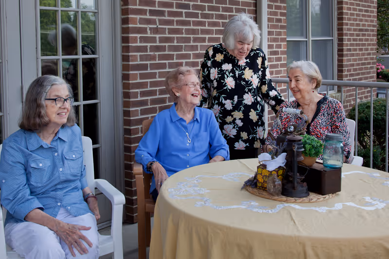 Four elderly women sitting and standing around a round table with a yellow tablecloth on a patio outside a brick building. The table has decorative items including a lantern, a small plant, and a jar. The women are smiling and appear to be enjoying each other's company.