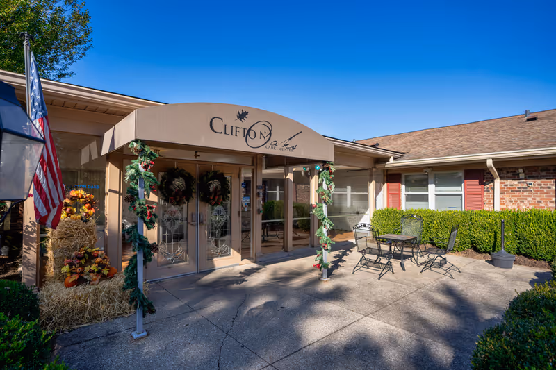 Entrance of Clifton Oaks Care Center decorated with holiday wreaths and garlands, an American flag on the left, and outdoor patio furniture on the right under a clear blue sky.