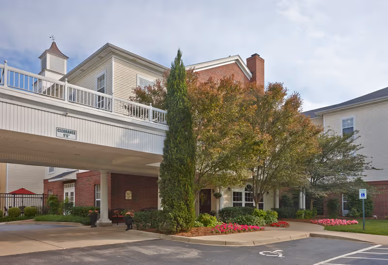 Exterior view of Belmont Village Senior Living St. Matthews showing a covered driveway entrance with clearance sign, surrounded by trees, shrubs, and flower beds. The building features a combination of brick and light-colored siding with multiple windows and a cupola on the roof.