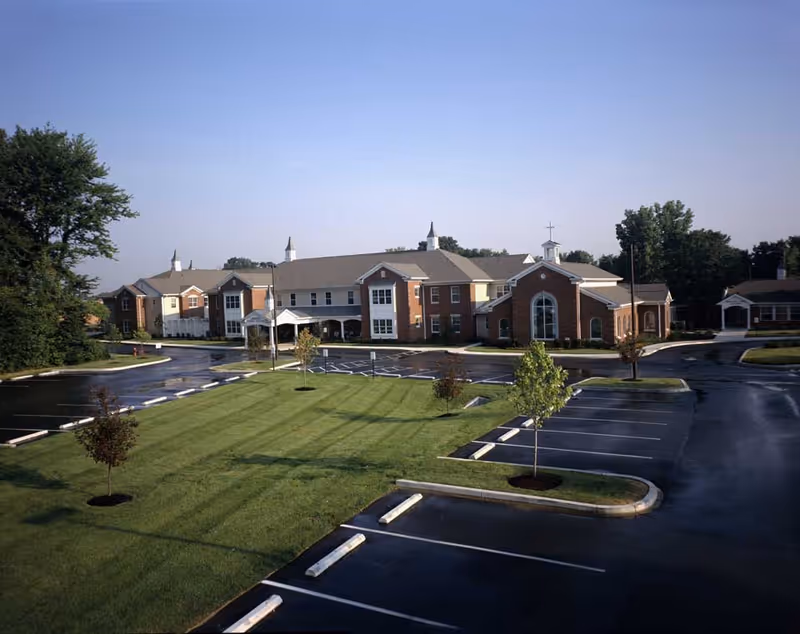 Exterior view of Franciscan Health Care Center showing a large brick building with multiple windows and steeples, surrounded by a well-maintained lawn and an empty parking lot with several small trees planted in landscaped islands.