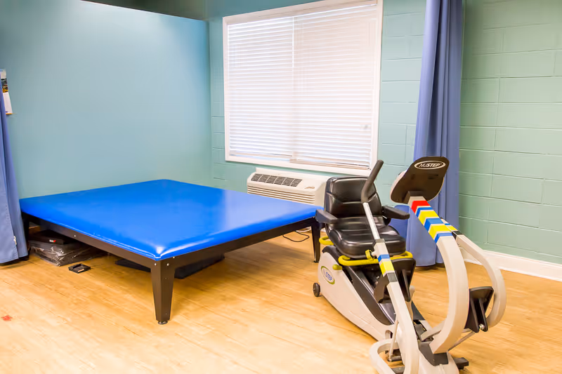 A room with light blue walls and wood flooring containing a large blue padded therapy table and a NuStep exercise machine positioned near a window with closed blinds.