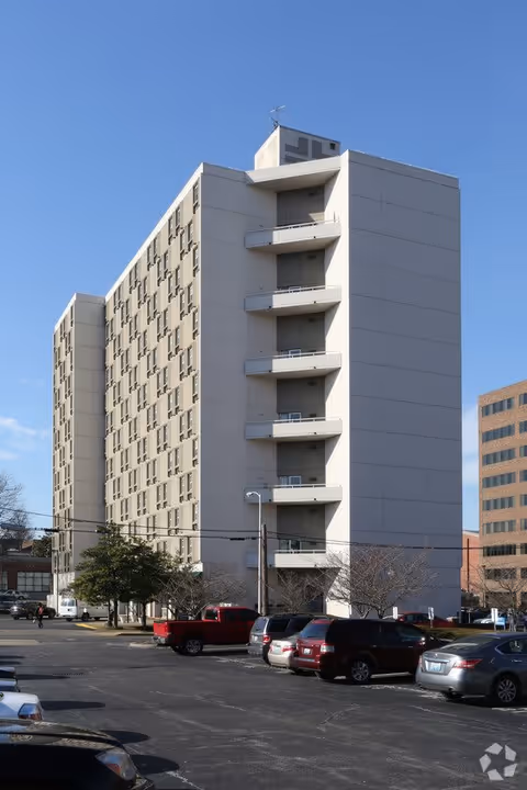 A tall, beige multi-story building with balconies on one side, surrounded by a parking lot with several parked cars under a clear blue sky.