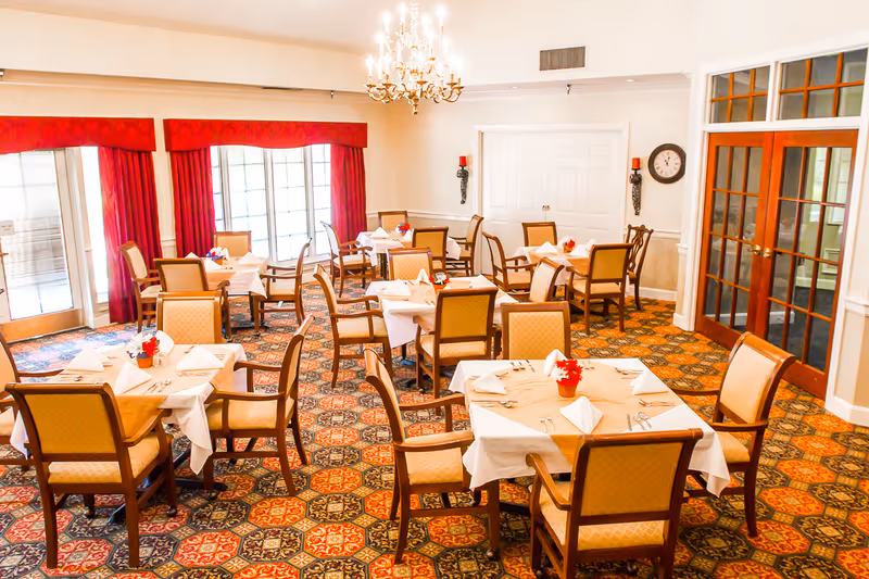 A bright and elegant dining room with multiple tables covered in white tablecloths, each set with napkins, silverware, and small red flower centerpieces. The room features patterned carpet, red curtains on large windows, a chandelier hanging from the ceiling, and wooden chairs with cushioned seats. There are double doors and a clock on the wall.