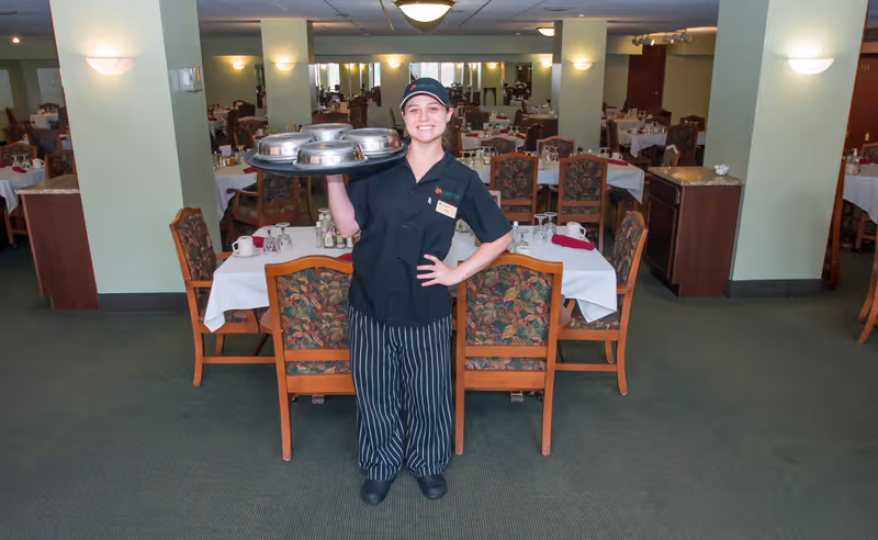 A smiling server wearing a black uniform and striped pants stands in a dining room holding a tray with covered dishes. The dining room has multiple tables set with white tablecloths, glasses, and napkins, and floral upholstered chairs. The room is well-lit with wall sconces and ceiling lights.