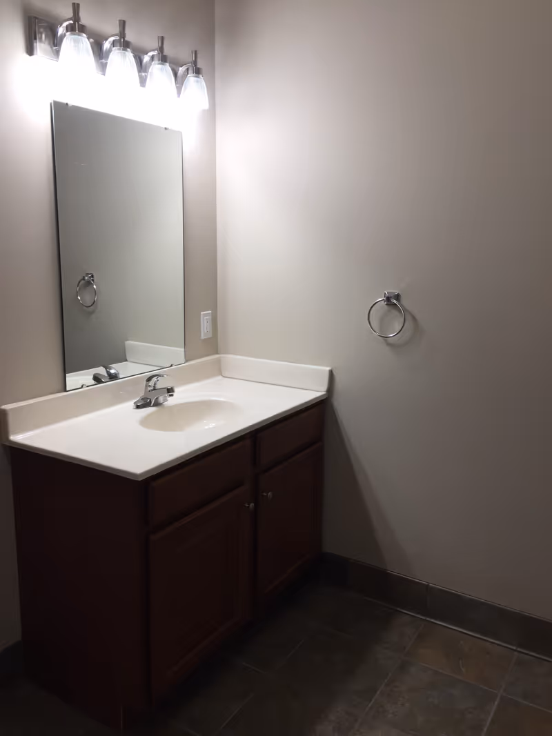 A bathroom vanity with a sink, mirror, overhead lights, and a towel ring on a neutral-colored wall.