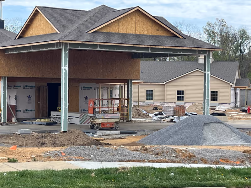 Partially built assisted living facility entrance with exposed sheathing, construction equipment, and gravel piles on site.