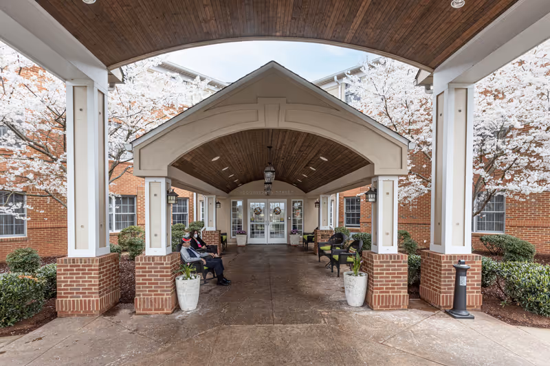 Covered entrance portico of a brick senior living building with benches, planters, blooming trees, and two people seated.