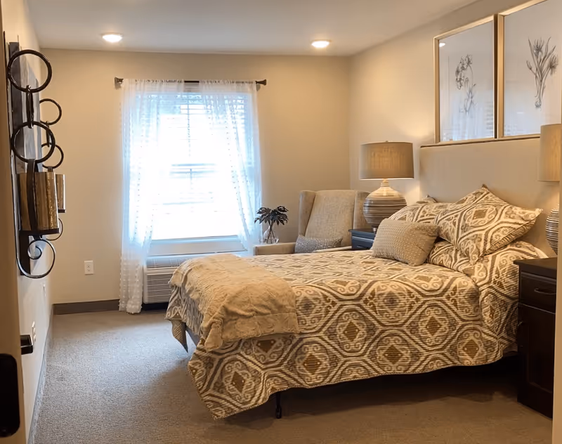 A cozy bedroom with a patterned bedspread on a bed, two matching pillows, and a beige throw blanket. There is a beige upholstered armchair next to the bed, two dark wooden nightstands with matching lamps, and two framed floral artworks hanging above the bed. A window with sheer white curtains allows natural light into the room.