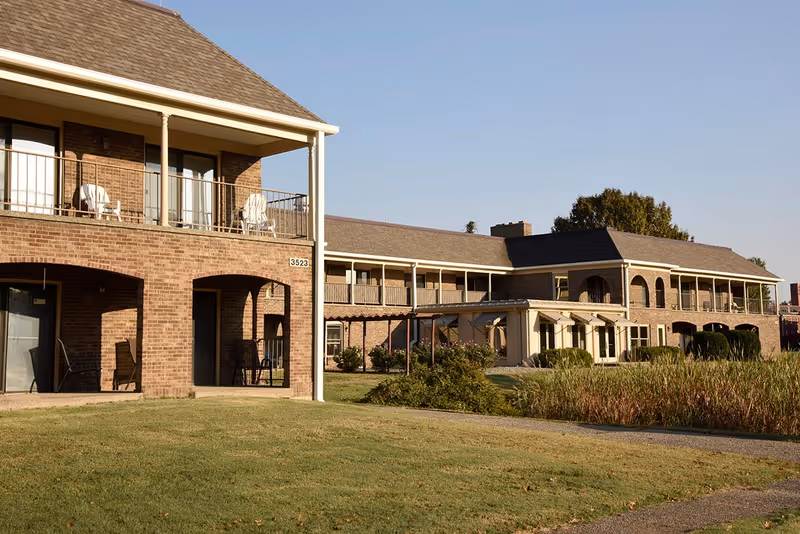 Exterior view of Twinbrook Assisted Living Apartments showing a two-story brick building with balconies and outdoor chairs, surrounded by grass and bushes under a clear sky.
