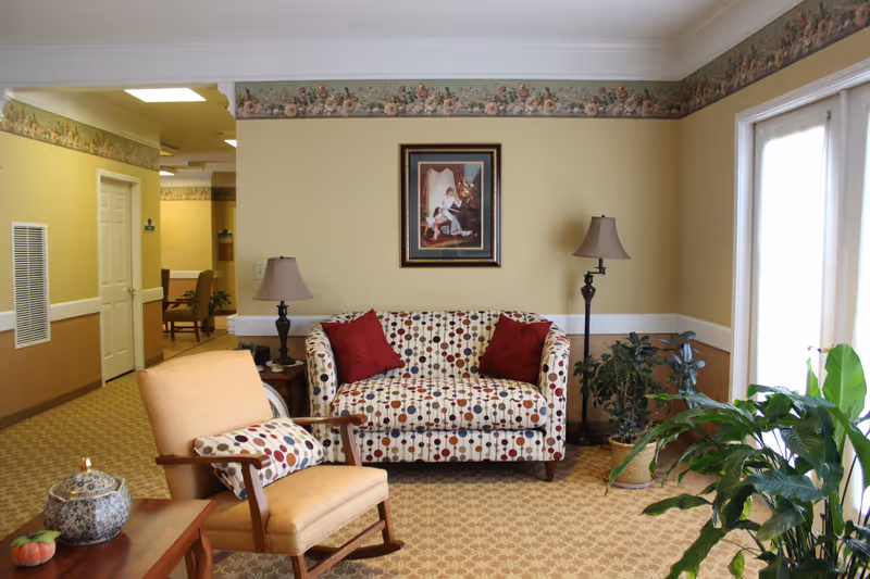 A cozy senior living facility common area with a patterned loveseat adorned with two red pillows, a beige armchair with a matching pillow, two lamps on either side of the loveseat, a framed painting on the wall, potted plants, and a table with a decorative ceramic container. The room has beige walls with floral wallpaper border near the ceiling and carpeted floor with a geometric pattern.