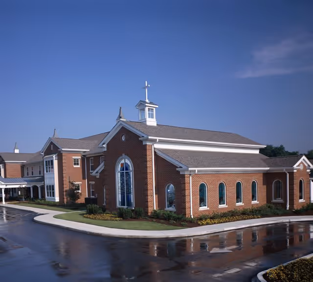 Brick chapel-style building with arched windows, a small cupola and cross, and an adjacent facility entrance under a blue sky.