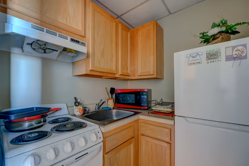 Compact kitchenette with an electric stove, sink, red microwave, white refrigerator and wooden cabinets.