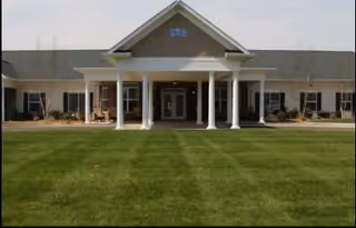 Front exterior view of a single-story senior living facility building with a covered entrance supported by white columns, large windows, and a well-maintained green lawn in front.