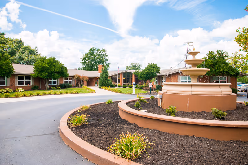 Exterior view of Fountain Circle Care & Rehabilitation Center showing a circular driveway with a tiered water fountain in the center, surrounded by landscaped plants and trees. The building is a single-story brick structure with large windows and an American flag near the entrance under a partly cloudy sky.