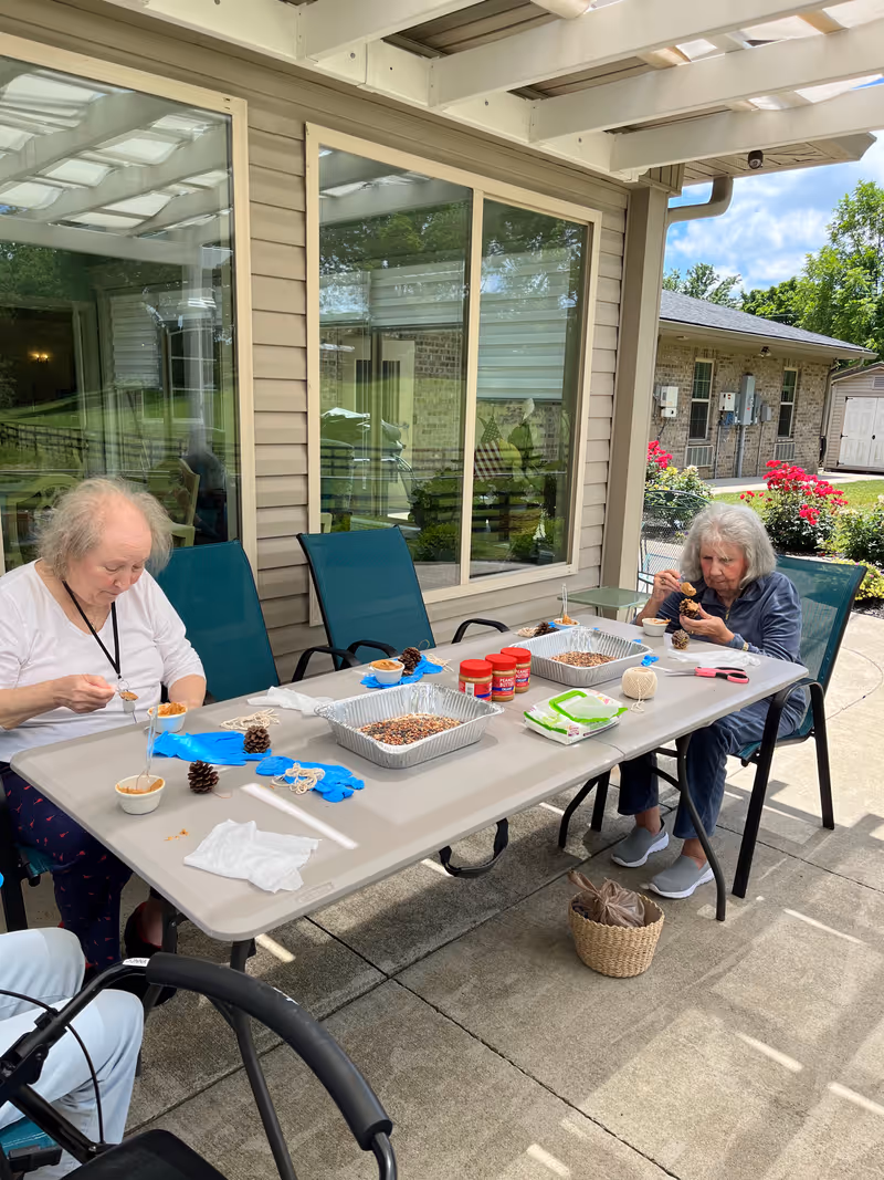 Two elderly women sitting at a table outdoors under a covered patio, engaging in a craft activity involving pine cones, peanut butter, and birdseed. The table has trays with birdseed, jars of peanut butter, string, gloves, and other craft supplies. The background shows a building with windows and a garden with flowers.