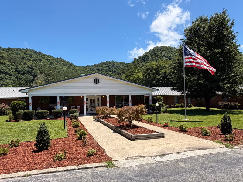 Single-story facility facade with white front, American flag, walkway, and landscaped entrance on a sunny day.