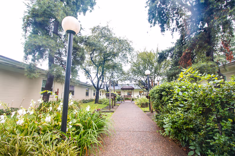 A landscaped courtyard walkway between single-story buildings with lamp posts, trees, and benches.