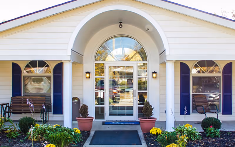 Front entrance of a building with a white exterior, an arched doorway with glass double doors, two potted plants on either side of the entrance, two windows with blue shutters, and benches on both sides of the entrance. There are also some flowers and greenery in front of the building.