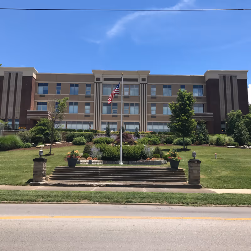 Front exterior view of a multi-story assisted living facility building with a flagpole displaying the American flag in the center of a landscaped lawn with trees, shrubs, and flower pots. The sky is clear and blue.