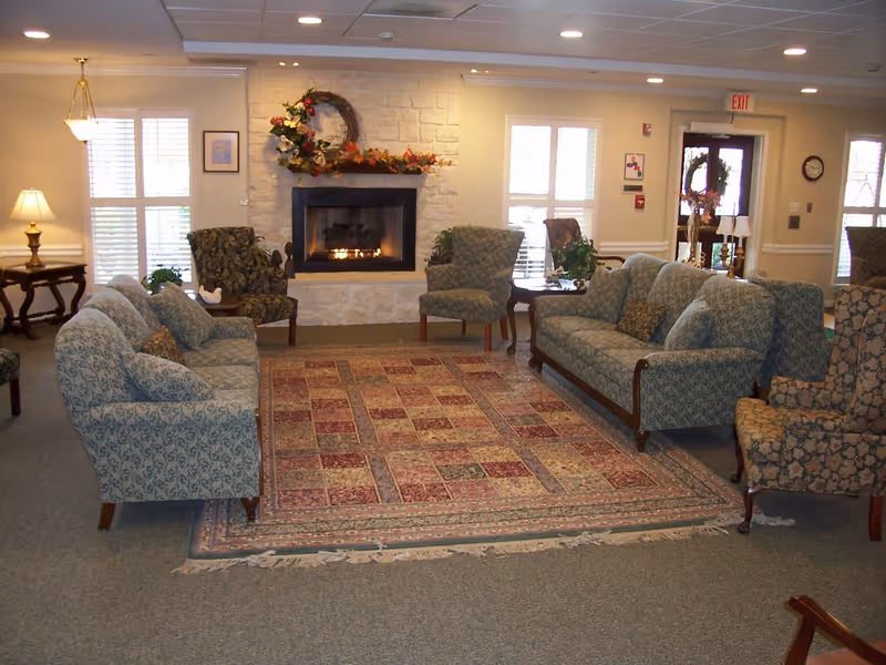 A cozy senior living facility common area with two patterned sofas and several armchairs arranged around a large decorative rug. A stone fireplace with a floral wreath and autumn decorations is centered on the far wall. The room is softly lit with table lamps and natural light coming through windows with white shutters. There is an exit door with a wreath and a clock on the wall.