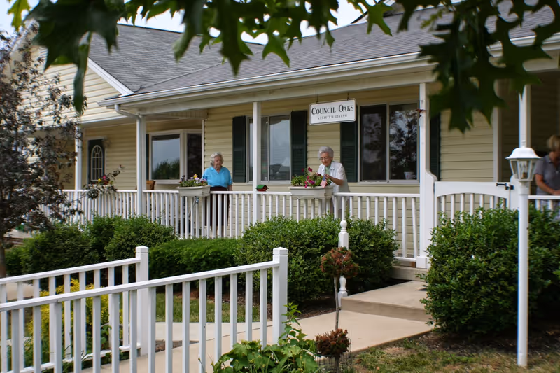 Front porch of a yellow assisted living building with white railing, a 'Council Oaks Assisted Living' sign, two elderly women tending plants on the porch, and shrubs in the foreground.