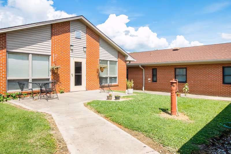 Sunlit brick courtyard and entrance of a senior living building with patio chairs, a concrete walkway, and a red hydrant post.