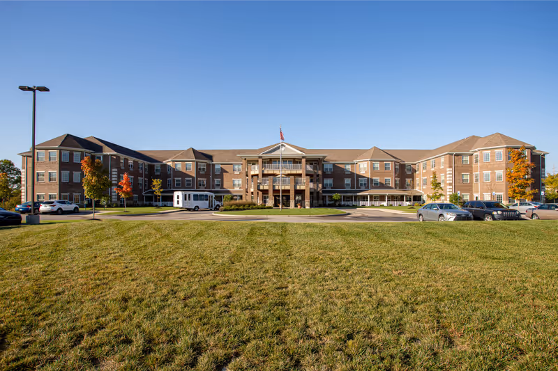 Front exterior view of Magnolia Springs Florence, a large three-story brick building with multiple windows, a central entrance with balconies, an American flag on a flagpole, a circular driveway, parked cars, and a well-maintained grassy lawn in the foreground under a clear blue sky.