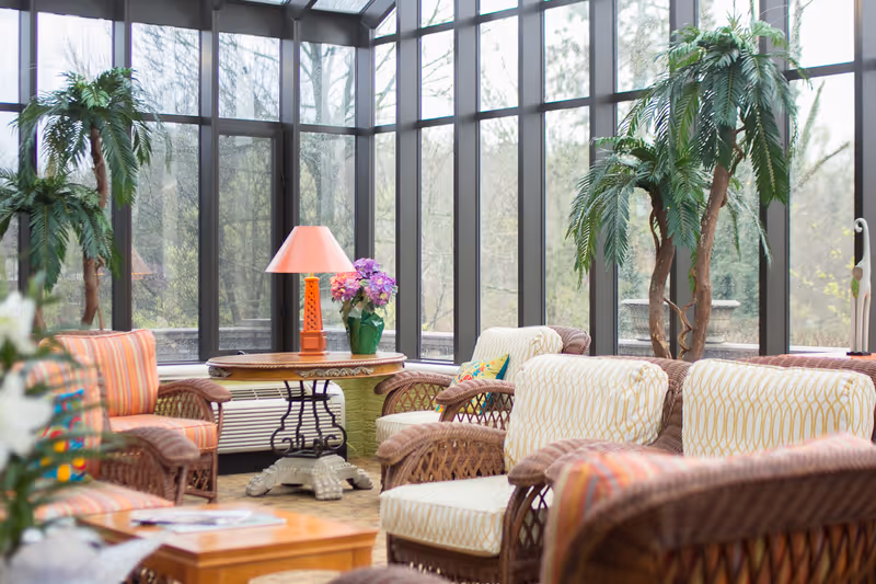 A bright sunroom with large floor-to-ceiling windows showing an outdoor view of trees. The room is furnished with wicker chairs and sofas with patterned cushions, a round wooden table with a pink lamp and a vase of flowers, and several potted plants.