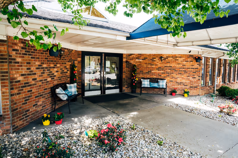 Covered brick entrance with double glass doors, benches, potted flowers and a rock garden under a canopy.