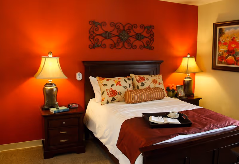 A cozy bedroom with a dark wooden bed frame and white bedding accented by floral and striped pillows. There are two bedside tables, each with a lamp, and a decorative metal wall art piece above the bed. A tray with a cup, glasses, and a book is placed on the bed. The walls are painted red and beige, and a colorful floral painting hangs on the beige wall.