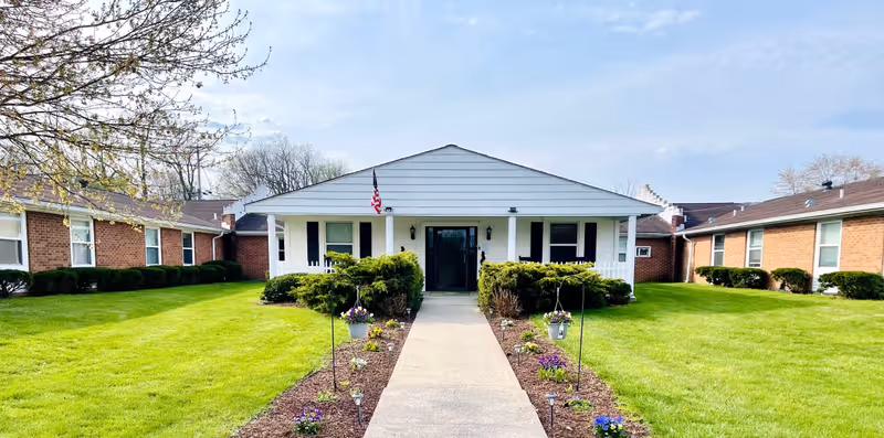 Front exterior view of a single-story nursing and rehabilitation center building with a white facade, black shutters, and an American flag near the entrance. A concrete walkway leads to the entrance, flanked by flower beds and green lawns on both sides. The building is surrounded by other brick buildings and trees under a partly cloudy sky.