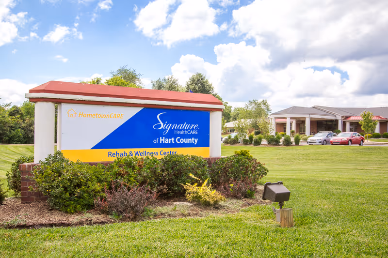 Entrance sign for Signature HealthCARE of Hart County Rehab & Wellness Center on a grassy lawn with the facility building and parked cars in the background.