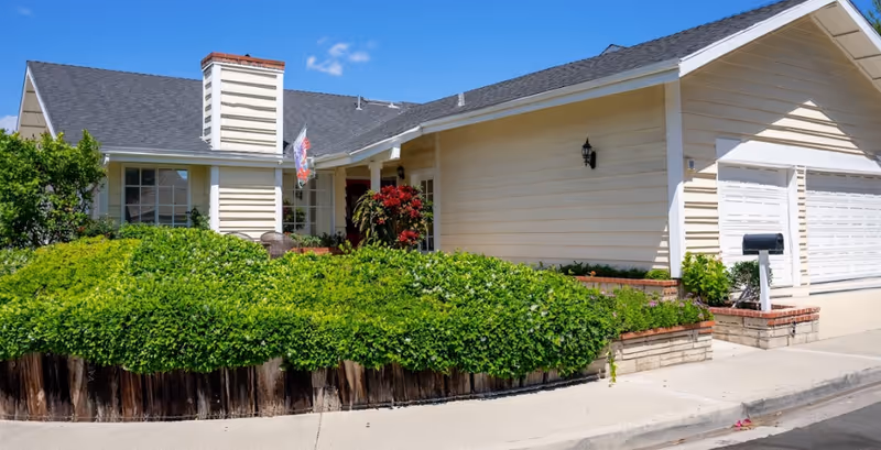 Single-story beige senior living facility with manicured hedges, a driveway, and a white garage.