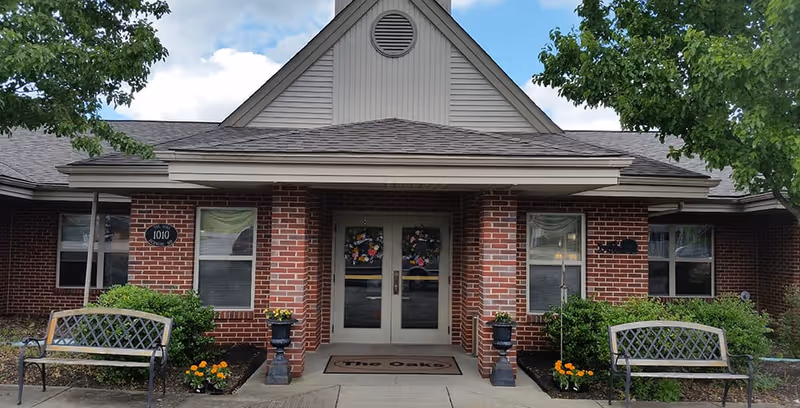 Brick-front entrance to The Oaks assisted living with double glass doors, a 'The Oaks' doormat, benches, planters and decorative wreaths.