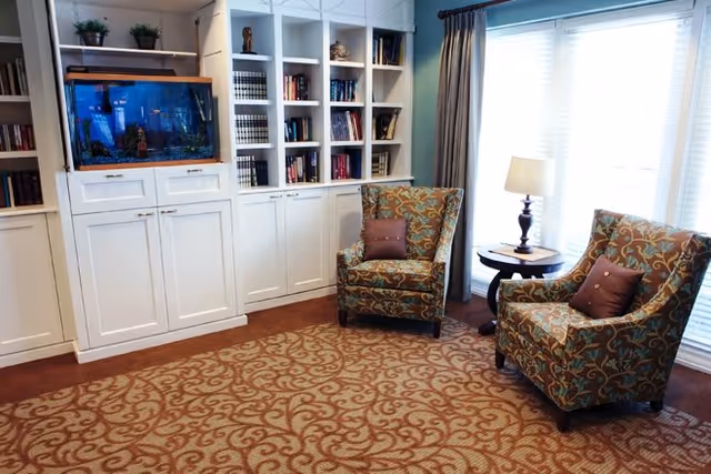 A cozy living room area with two patterned armchairs each with a small pillow, a round side table with a lamp, large windows with curtains letting in natural light, a built-in white bookshelf filled with books and decorative items, and a large fish tank on the left side.