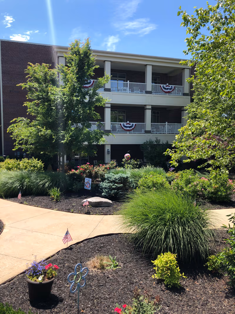 Outdoor garden area with various green plants, shrubs, and flowers in front of a three-story brick building with balconies decorated with red, white, and blue bunting. A paved walkway curves through the garden, and small American flags are placed along the path.
