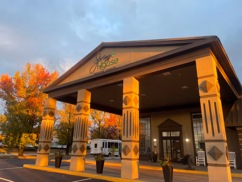 Front entrance of The Spring House senior living building with a covered porte-cochere and decorative columns lit by warm sunset light.
