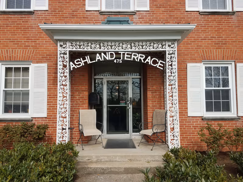 Brick building entrance with a white decorative awning reading ASHLAND TERRACE and two chairs flanking the doorway.