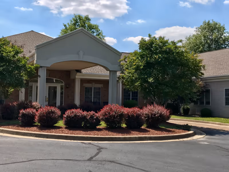 Front entrance of a single-story building with a covered portico, landscaped shrubs and trees under a blue sky.