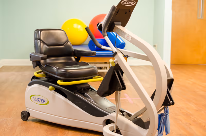 A NuStep recumbent cross trainer exercise machine with a black seat and yellow handrails is placed on a wooden floor. In the background, there are colorful exercise balls and a padded bench against a light green wall.
