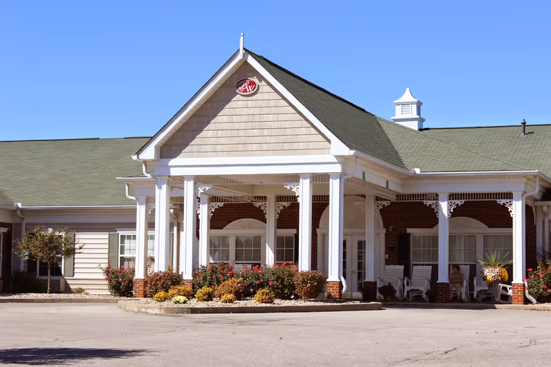 Exterior front entrance of a senior living building with a covered porch supported by white columns, rocking chairs, and landscaped flower beds.