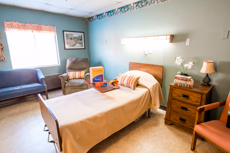 A well-lit room in a senior living facility featuring a single bed with a beige blanket and an orange-striped pillow. Next to the bed is a wooden nightstand with a lamp, books, and a small plant. There is a brown armchair with an orange cushion, a blue patterned loveseat, and a recliner chair near a window with blinds and a peach-colored valance. A framed picture hangs on the green wall above the recliner. A welcome book and a small flower arrangement are placed on a tray over the bed.
