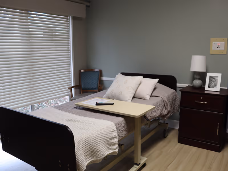 A small bedroom with a single hospital-style bed covered with a beige quilt and pillows. A beige blanket is draped over the foot of the bed. There is a light-colored overbed table with a remote control and papers on it. Next to the bed is a dark wooden nightstand with a lamp and a framed photo. A wooden chair with blue upholstery is placed near a window with closed white blinds. The walls are painted a muted green color and the floor is light wood.