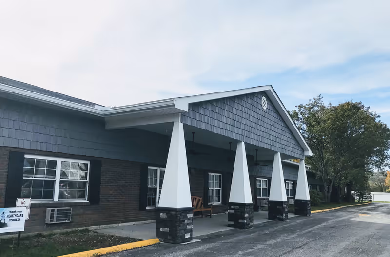Exterior view of Wurtland Nursing & Rehabilitation facility showing a covered entrance with four white pillars, brick walls, windows with black shutters, and a tree on the right side under a partly cloudy sky.