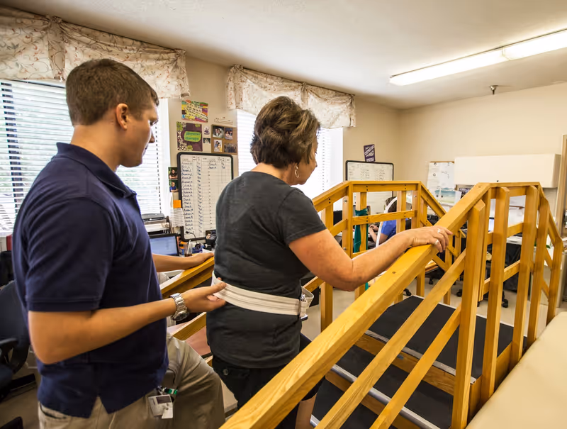A caregiver assists a woman walking up a wooden ramp inside a room with windows and whiteboards on the walls. The woman holds the handrail while the caregiver supports her with a gait belt around her waist.