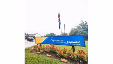 Exterior view of a landscaped entrance sign reading 'Signature HealthCARE at Colonial Rehab & Wellness Center' with flags and part of the facility visible behind it.
