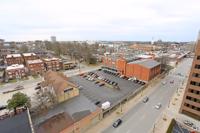 Aerial view of an urban area showing a parking lot with several cars, surrounding buildings including a large brick building, residential houses, and a main road with a few vehicles driving on it under a cloudy sky.