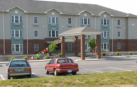 Exterior view of a three-story senior living facility building with a covered entrance supported by brick columns. Two cars are parked in the parking lot in front of the building, and colorful triangular flags are strung along the entrance walkway.